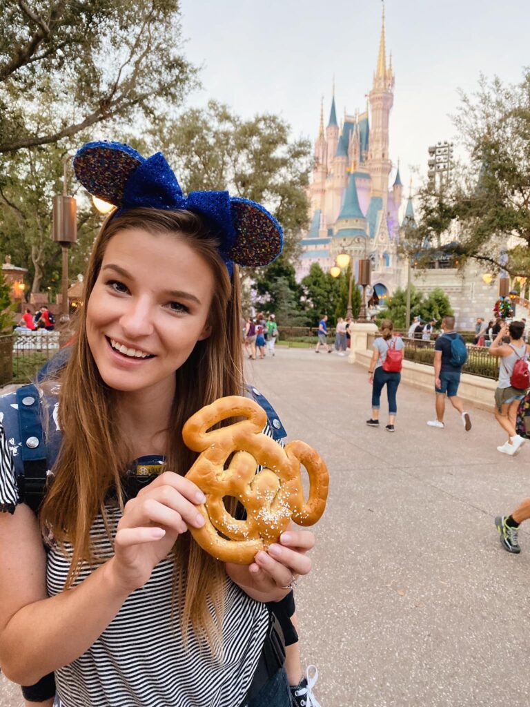 Woman holding a lightly salted Mickey Mouse pretzel standing in front of the Magic kingdom Cinderella Castle